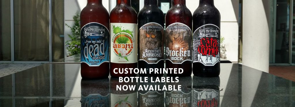 Custom Bottle labels printed for Bury Me Brewing in Florida placed on the bottles and all the bottles lined up on table with text stating custom printed bottle labels now available